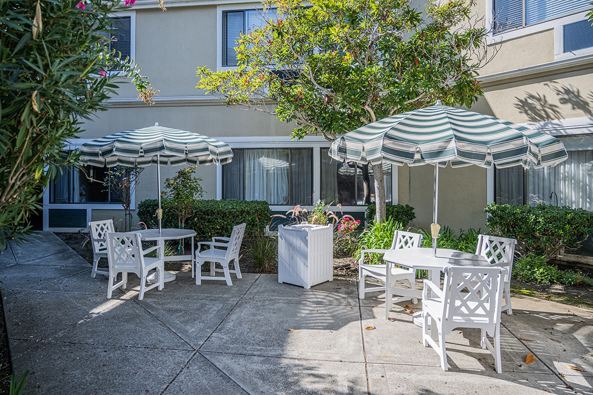 Outside courtyard with two sets of white tables and chairs, striped umbrellas and greenery